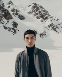 Asian male model wearing a grey zip jacket and black turtleneck against a snowy mountain range backdrop