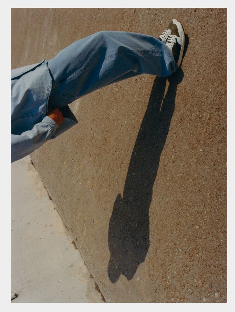 Person in blue denim jacket and sneakers sitting on concrete ground with a shadow of a hand on a sunny day outdoors.