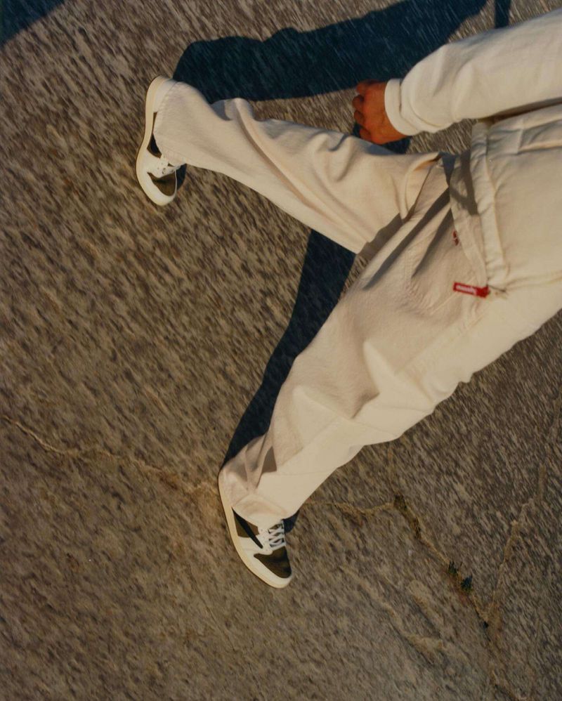 Person in white pants and black-and-white sneakers stands on mottled grey surface with sharp shadow. Fashion and style concept.