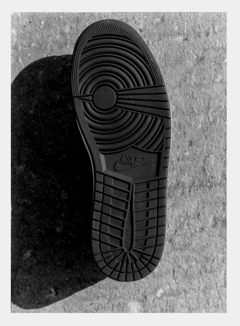 Close-up of the textured black sole of a Nike sneaker, with the Nike logo, set against a gray concrete background with a prominent shadow.