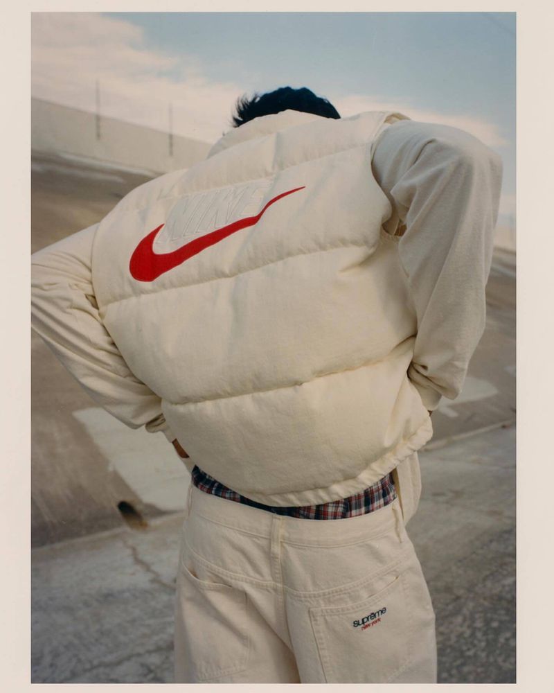 Person wearing a white puffer vest with a large red Nike swoosh on the back, off-white jeans with a Supreme logo, and plaid shirt.