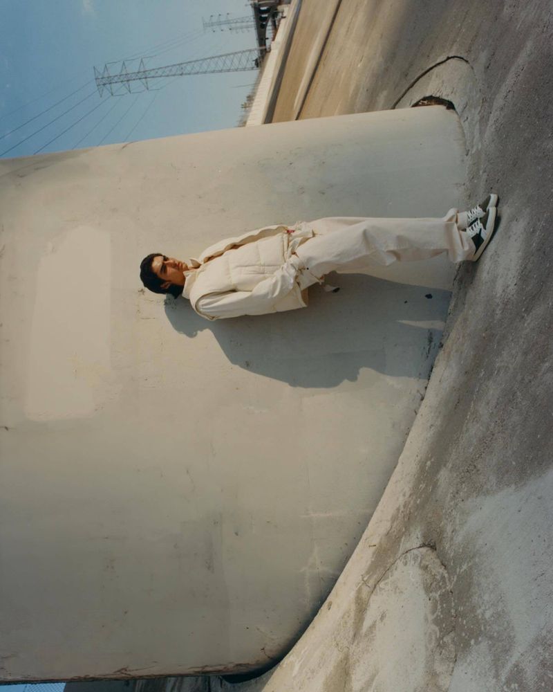 A person in white clothes poses next to a concrete structure with power lines visible in the background on a sunny day outdoors.