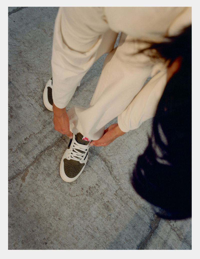 Overhead shot of a person tying the laces of a dark-green-and-white retro sneaker while wearing beige pants on a grey surface.