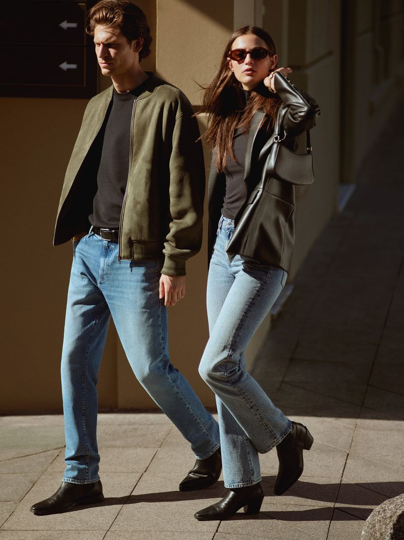 A stylish couple walks outside wearing fashionable jeans, jackets, and boots, showcasing modern street style and casual chic in daylight.