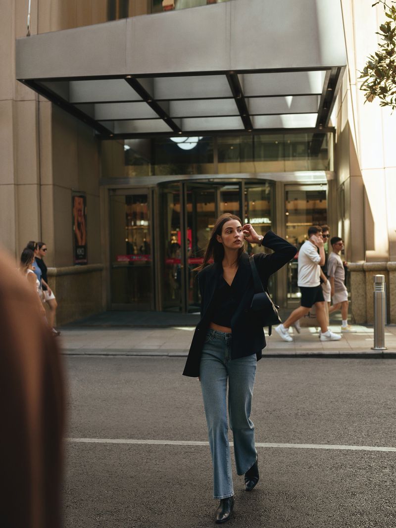 Woman in blazer and jeans walks along a city street with pedestrians and a building with revolving doors in the background.
