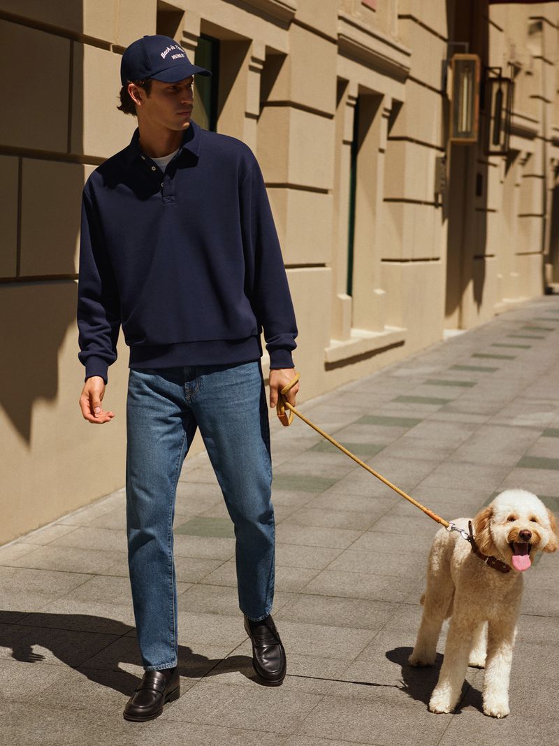 Man walking a dog on a leash in an urban setting, wearing a navy blue sweatshirt, jeans, and a baseball cap on a sunny day.