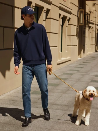 Man walking a dog on a leash in an urban setting, wearing a navy blue sweatshirt, jeans, and a baseball cap on a sunny day.