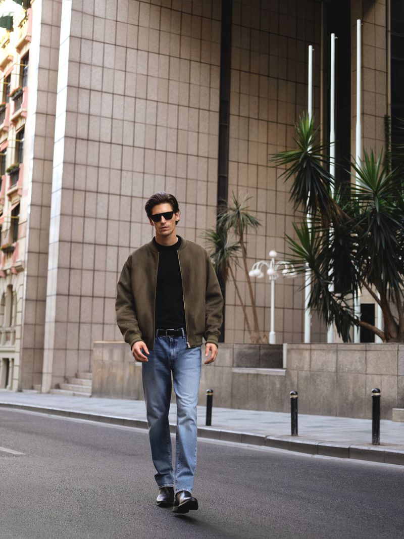 A stylish man walks on a city street in an olive bomber jacket, black shirt, jeans, and sunglasses with tall buildings behind him.