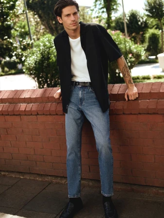 Man in casual blazer, jeans, and black shoes leaning against brick wall outdoors with trees and greenery behind him in daylight.