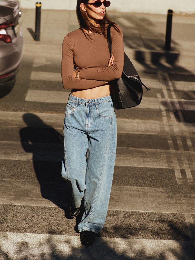 Fashion shot of a woman wearing a long-sleeved brown top and oversized blue jeans walking across a crosswalk on a sunny day.