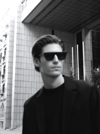 Black and white portrait of an adult man in sunglasses and blazer outdoors in an urban environment, with city buildings in the background.
