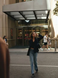 Model in dark jacket and blue jeans walking on city street in front of a modern hotel entrance
