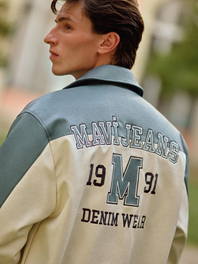 Man with dark hair poses in a blue, cream, and black jacket with 'Mavi Jeans' and 'Denim Wear' printed on the back outdoors.