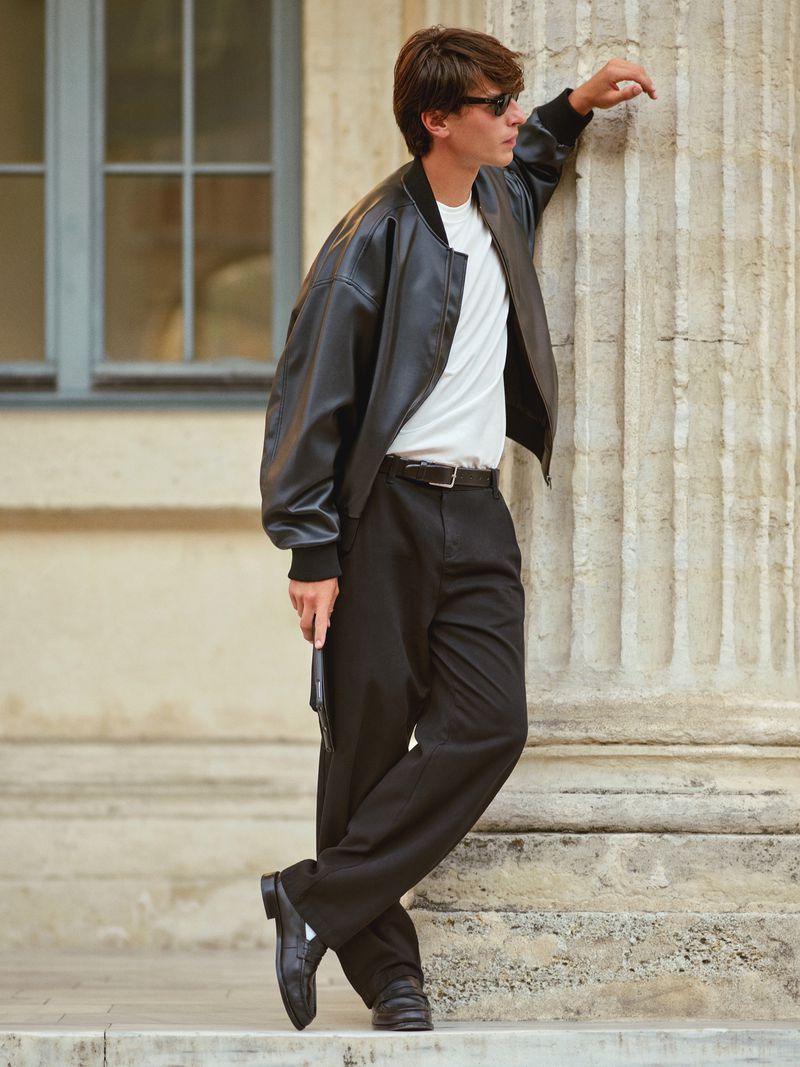 Young man in sunglasses, black bomber jacket, white t-shirt, and black pants leaning against a fluted column in an outdoor setting.