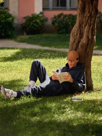 A young man with a baseball cap is relaxing on the grass and reading a book near a tree on a sunny day in an outdoor setting.