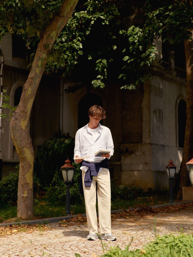 Full shot of a young adult male standing outside in casual clothes, looking down at a handheld device in a park-like setting.
