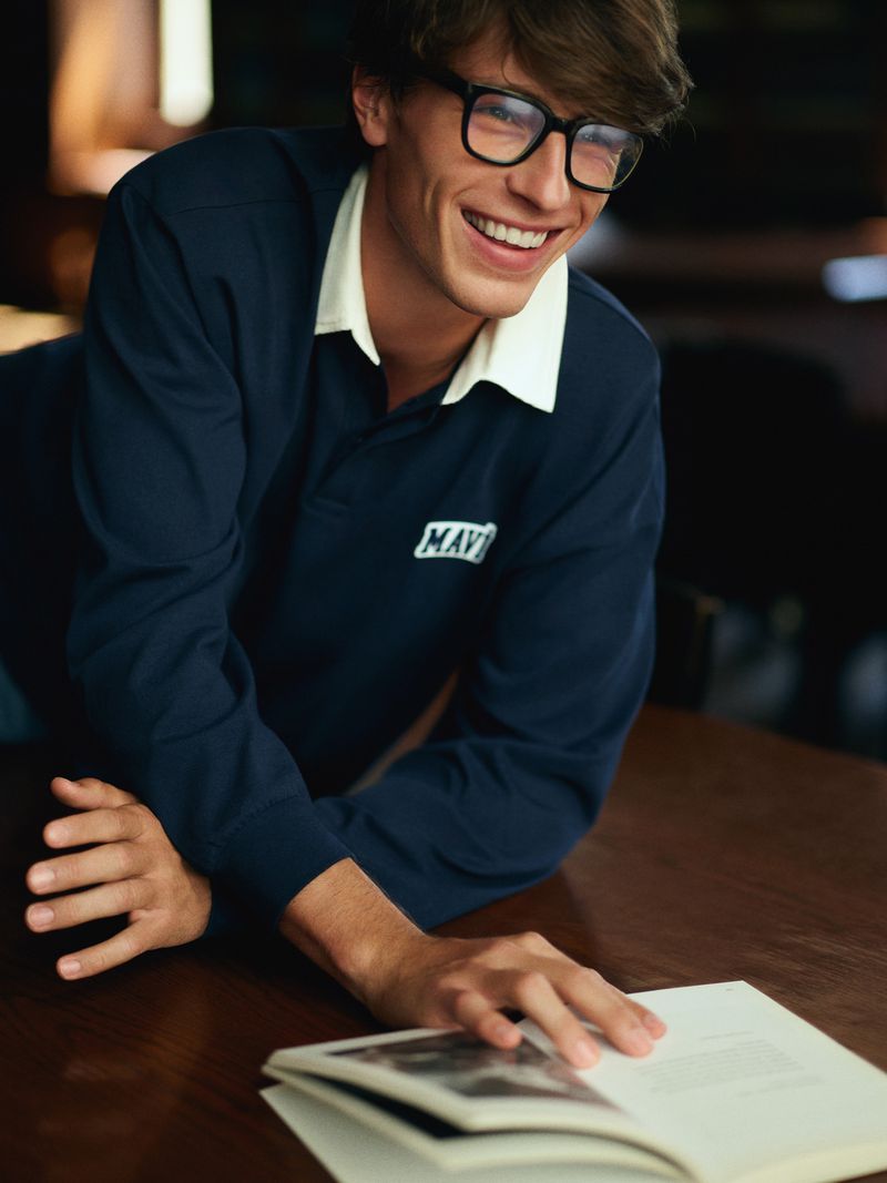 Smiling adult male wearing glasses and a navy long-sleeved shirt with a white collar, leaning on a dark wood table reading a book.