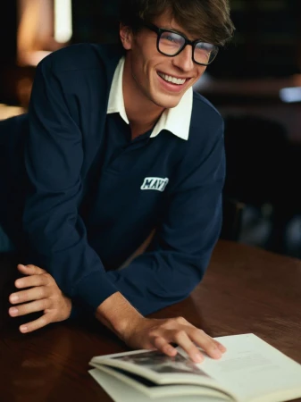 Smiling adult male wearing glasses and a navy long-sleeved shirt with a white collar, leaning on a dark wood table reading a book.