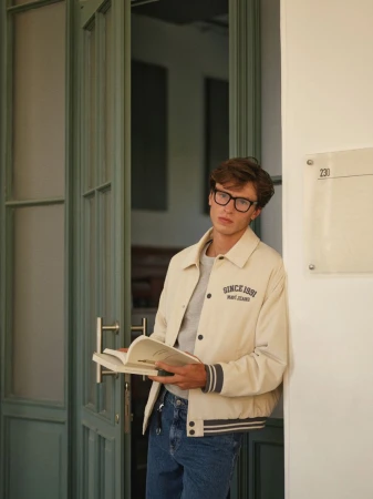 Young man in glasses leans against a doorway, holding a book and wearing a cream-colored jacket with jeans in natural light.