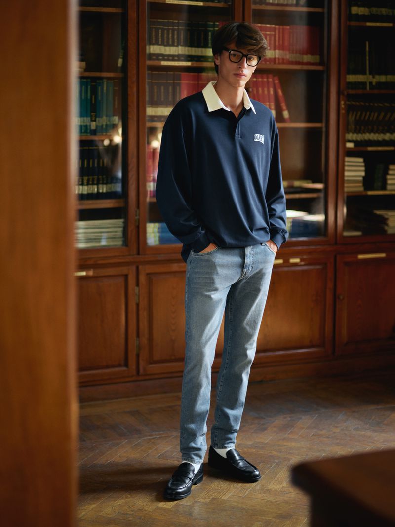 Male model in glasses wearing a navy polo shirt, light blue jeans, and black loafers standing indoors with a library backdrop.