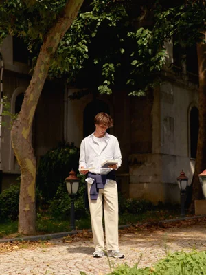 Young man in a white shirt and cream trousers reading a book outdoors near trees and an old building.