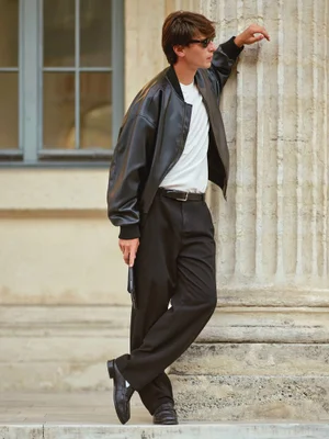 Model Clément Laguardia wearing a black leather jacket, white t-shirt, and black trousers while leaning against a stone column.