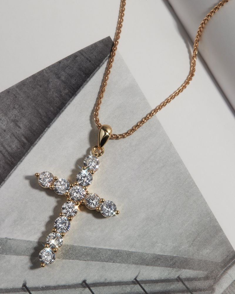 Close-up of a gold cross pendant necklace with clear gems, set against a backdrop of white and grey geometric patterns.