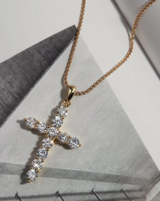 Close-up of a gold cross pendant necklace with clear gems, set against a backdrop of white and grey geometric patterns.