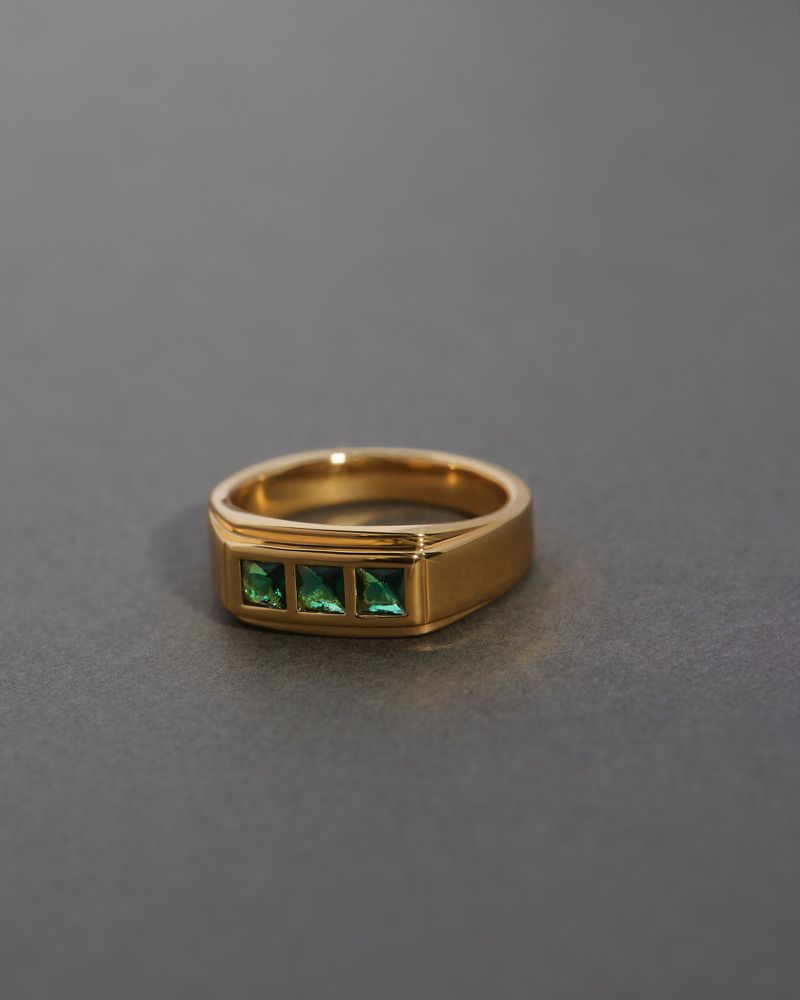 Gold ring with three green gemstone details on a grey surface in a studio product shot for jewelry product catalog and online marketing.