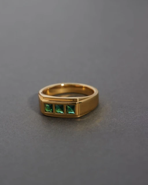 Gold ring with three green gemstone details on a grey surface in a studio product shot for jewelry product catalog and online marketing.