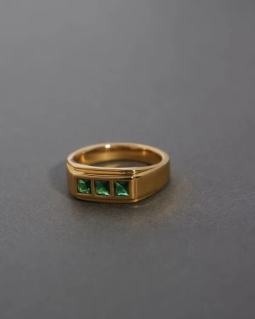 Gold ring with three green gemstone details on a grey surface in a studio product shot for jewelry product catalog and online marketing.