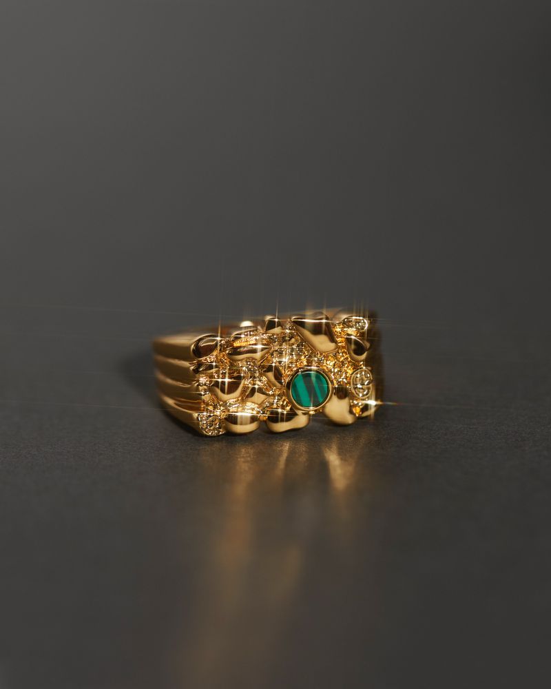 Close-up of a gold nugget ring with a green stone, casting a small shadow on a dark gray surface, showcasing jewelry and texture.
