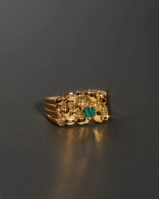 Close-up of a gold nugget ring with a green stone, casting a small shadow on a dark gray surface, showcasing jewelry and texture.