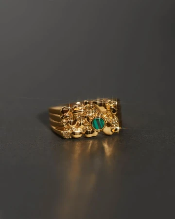 Close-up of a gold nugget ring with a green stone, casting a small shadow on a dark gray surface, showcasing jewelry and texture.