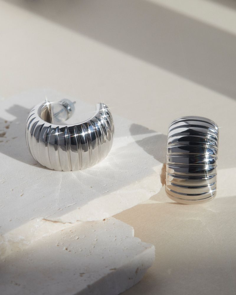 Studio shot of two modern silver hoop earrings with ridged texture on a reflective surface and a neutral background.