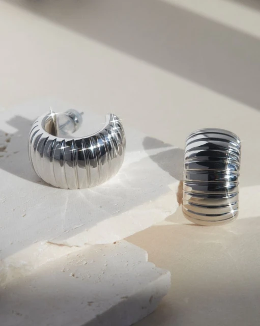 Studio shot of two modern silver hoop earrings with ridged texture on a reflective surface and a neutral background.