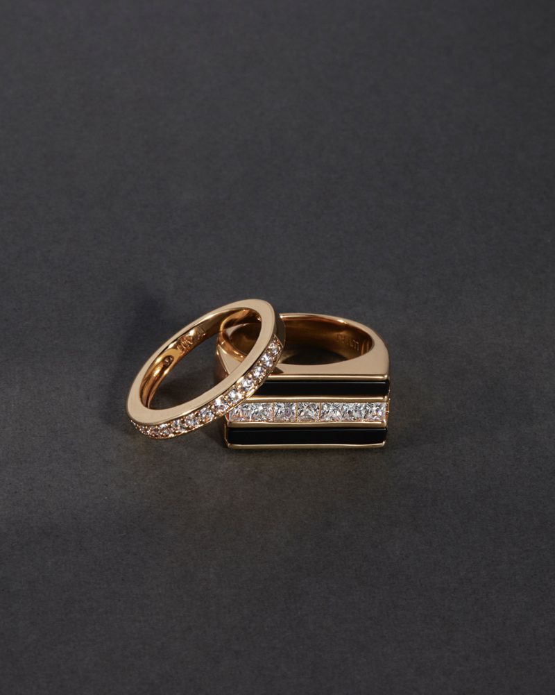 Close-up of two gold rings, one with black enamel and diamond accents, on a seamless dark gray background. Jewelry product shot.