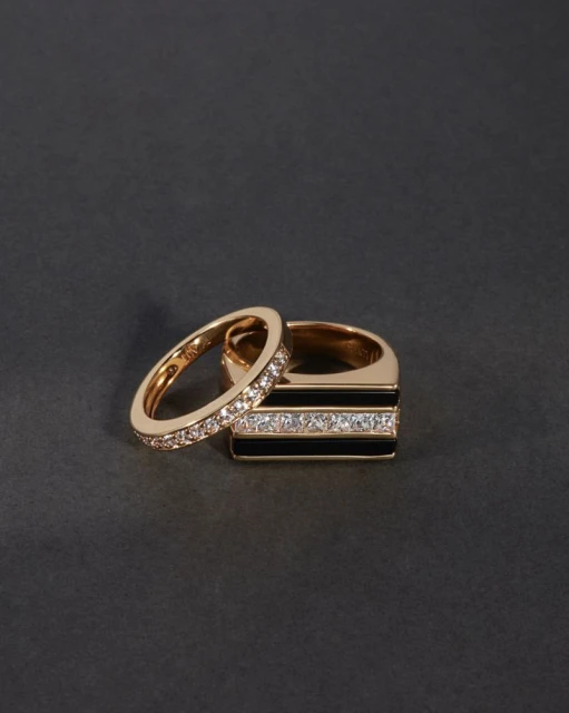 Close-up of two gold rings, one with black enamel and diamond accents, on a seamless dark gray background. Jewelry product shot.