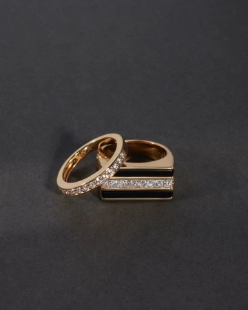 Close-up of two gold rings, one with black enamel and diamond accents, on a seamless dark gray background. Jewelry product shot.