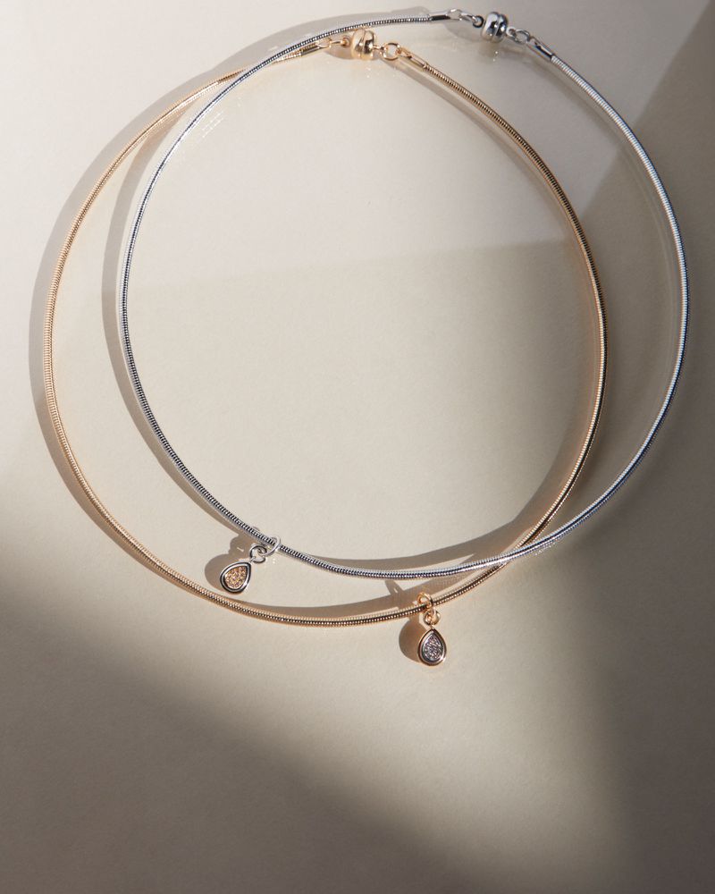 Two elegant gold and silver necklaces with teardrop pendants are displayed on a neutral surface with soft lighting creating shadows.
