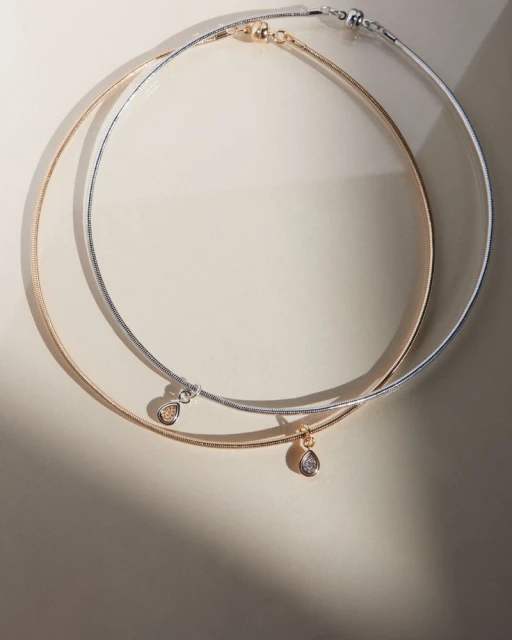 Two elegant gold and silver necklaces with teardrop pendants are displayed on a neutral surface with soft lighting creating shadows.