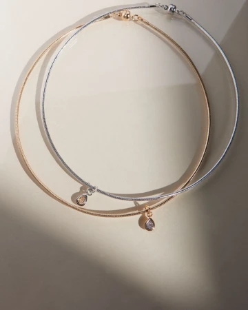 Two elegant gold and silver necklaces with teardrop pendants are displayed on a neutral surface with soft lighting creating shadows.