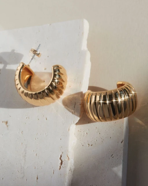 Two golden, ribbed hoop earrings sit on a textured white surface in a close-up shot showcasing minimalist fashion jewelry.