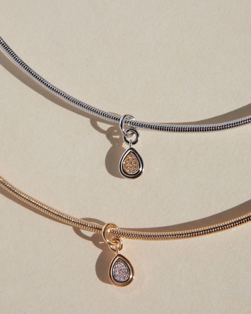 Close-up of two necklaces, one silver and one gold, with teardrop pendants against a soft beige background. Jewelry advertisement.