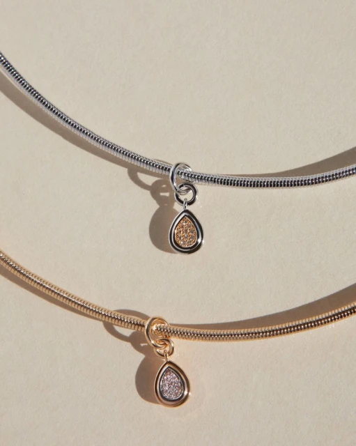 Close-up of two necklaces, one silver and one gold, with teardrop pendants against a soft beige background. Jewelry advertisement.
