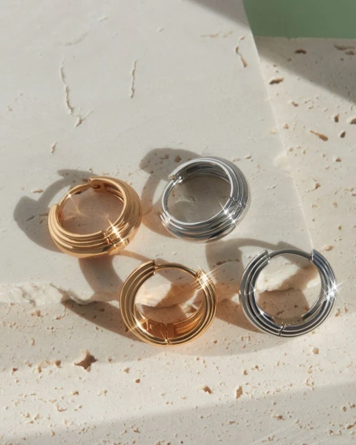 Four hoop earrings, two gold and two silver, arranged on a textured cream surface in a bright, even light, jewelry close-up.