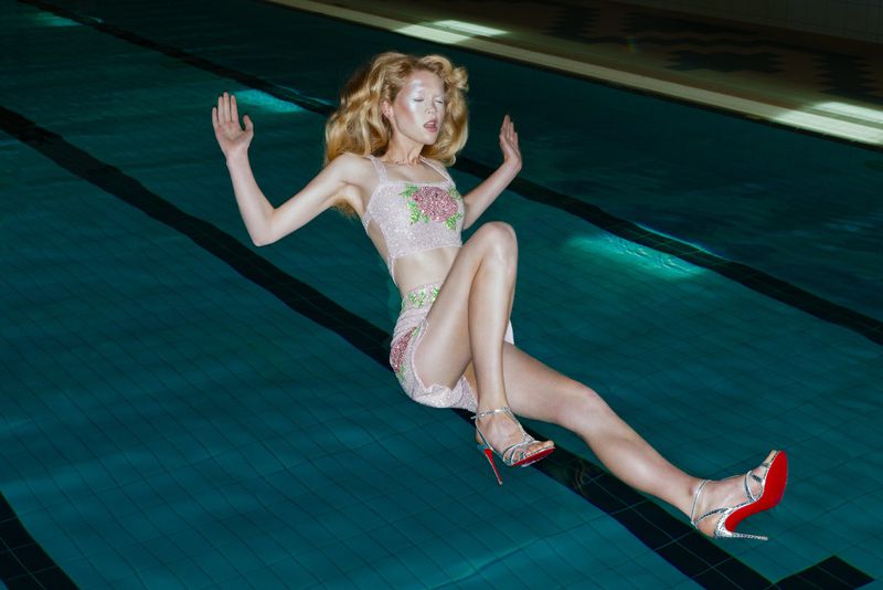Adult woman in beaded top and skirt poses in a swimming pool with a black lane marker, wearing high heels with red soles.