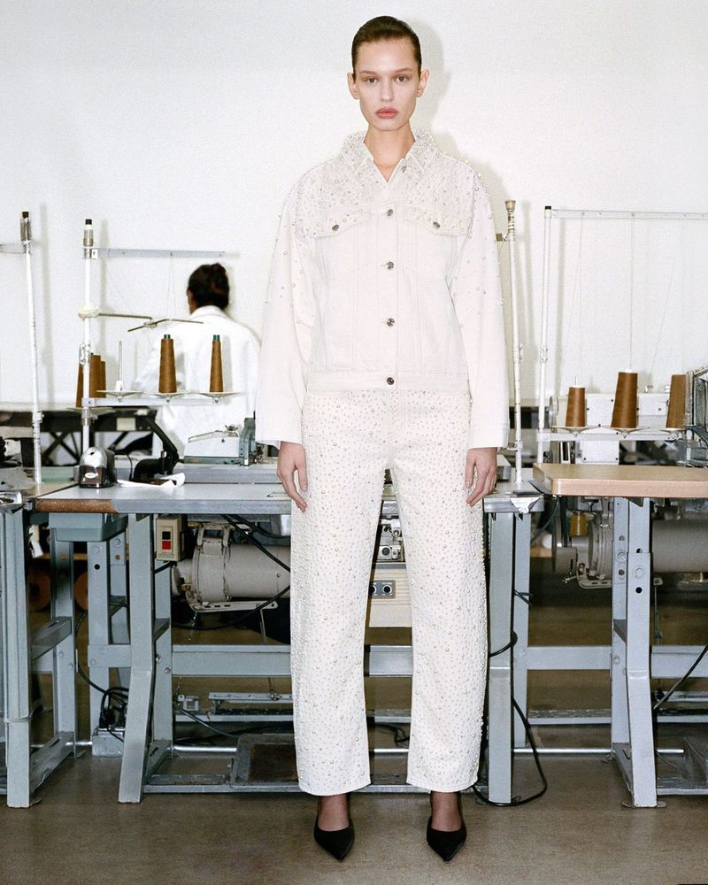 Fashion model posing indoors in a cream-colored, embellished jacket and pants with black heels in a sewing factory backdrop.