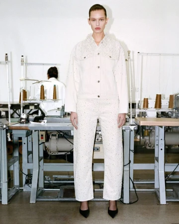 Fashion model posing indoors in a cream-colored, embellished jacket and pants with black heels in a sewing factory backdrop.