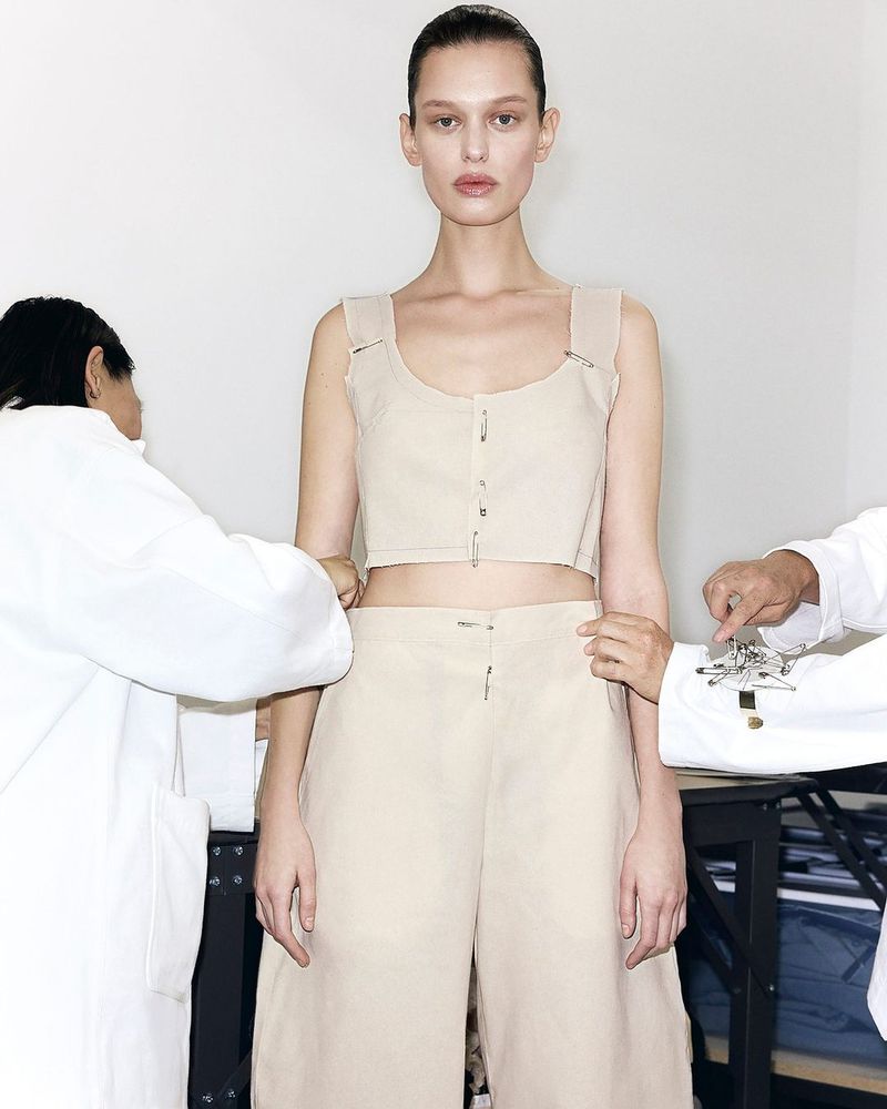 A model poses in a cropped top and wide-legged pants while being fitted by two unseen tailors in white lab coats.